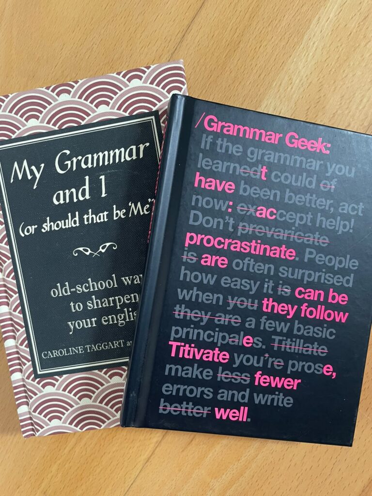 Two books lying fanned out on a wooden floor. The bottom book is entitled "My Grammar and I (or should that be 'Me')" by Caroline Taggart and J.A. Wines. The top book is "Grammar Geek" by Susanna Geoghegan, although the author's name is not visible on the cover.