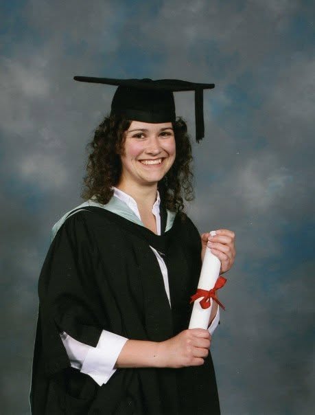 Official graduation picture from Lisa's Master's graduation ceremony. Lisa is wearing a black graduation robe with a light teal hood over a white shirt. She has a black graduation cap on her head and is smiling. In her hands she is holding a white roll of paper tied with a red ribbon.