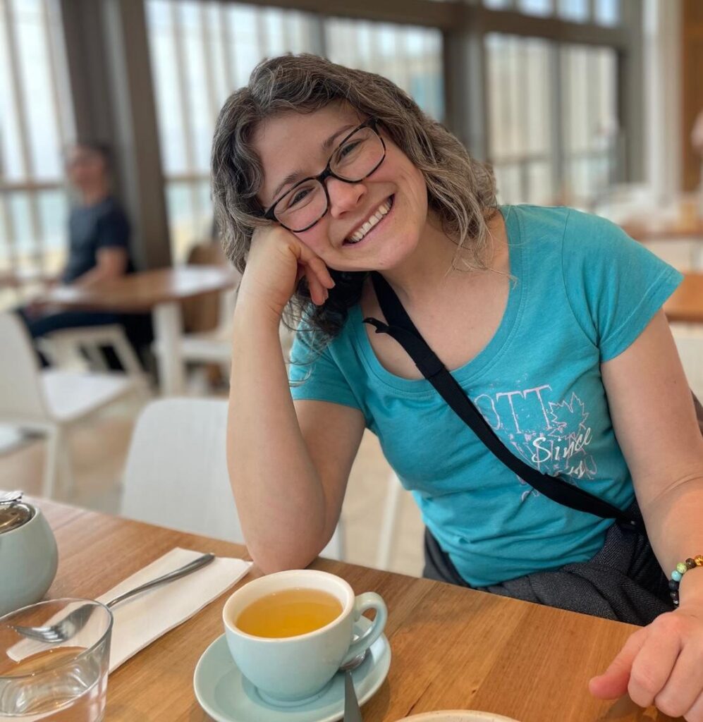 Lisa smiling directly into the camera, with her head tilted to one side and resting on her right hand. Her right elbow is leaning on the table in front of her. There is a cup of jasmine tea on the table in front of her, too, and a glass of water just behind that. Lisa is wearing a turquoise T-shirt. The background of the photo is blurred, but you can see that she is sitting in a spacious cafe with white plastic chairs at wooden tables. One wall of the room is made up of floor-to ceiling windows, though which you can just make out the azure blue of the sea.